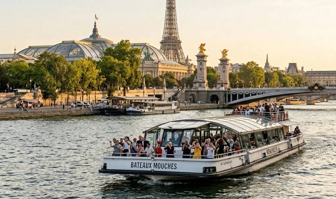 Ein Panoramablick auf Paris von der Seine aus im warmen Licht des späten Nachmittags. Ein Ausflugsboot mit dem Namen 'BATEAUX MOUCHES' in schwarzen Buchstaben an der Seite fährt mit Passagieren auf dem Fluss. Im Hintergrund erhebt sich der Eiffelturm über die Stadtsilhouette, flankiert vom Grand Palais links und der Pont Alexandre III mit seinen goldenen Statuen in der Mitte rechts. Das Ufer ist belebt.