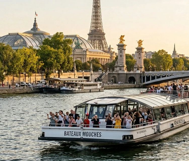 午後遅くの暖かい光の中でセーヌ川から見たパリのパノラマビュー。船体に黒い文字で「BATEAUX MOUCHES」と書かれた遊覧船が乗客を乗せて川を航行しています。背景にはエッフェル塔が都市の景観の上にそびえ、左にはグラン・パレ、中央右には金色の彫像があるアレクサンドル3世橋が見えます。岸辺は活気に満ちています。