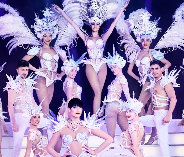 Dinner show table with champagne facing an illuminated Parisian cabaret stage with feathered dancers